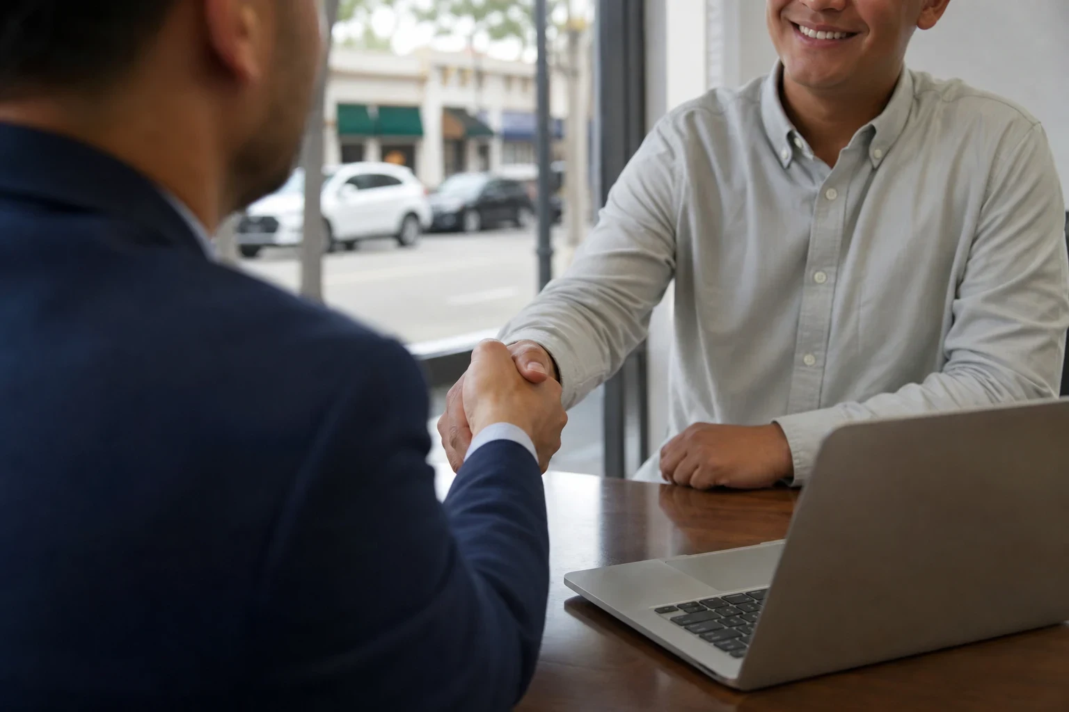 Over-the-shoulder view of an agent in a navy jacket shaking hands with a smiling client across a wooden desk with a laptop, a main street visible through the window