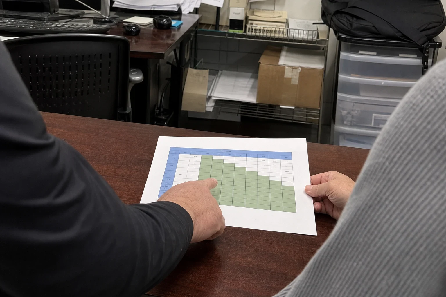 Over-the-shoulder view of two people at a wooden desk in a cluttered back office, one pointing at a printed coverage chart with blue and green table cells